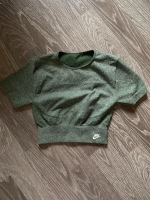 Grönt träningsset från Nike - Snyggt grönt träningsset från Nike med croppad t-shirt och matchande tights. Setet har ribbade detaljer och logga på både överdel och byxa. Perfekt för gymmet eller löprundan. Materialet är stretchigt och följsamt för maximal komfort. De är push up i rumpan därför ser de så skrynkligt ut på bilden ! Hela settet gick på 1700kr original pris. 
