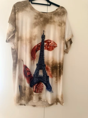 T-shirt med Eiffeltorn och fjädrar - Unik t-shirt med tryck av Eiffeltornet i mörkblått och röda fjädrar, dekorerad med små glittriga detaljer. T-shirten har korta ärmar och en lös passform. Färgerna går i beige, brun och vitt med en konstnärlig touch. Perfekt för dig som gillar Paris-vibbar!