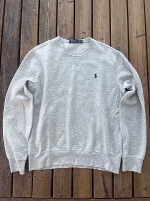 Grå sweatshirt från Polo Ralph Lauren - Klassisk grå sweatshirt från Polo Ralph Lauren med rund halsringning och broderad marinblå logga på bröstet. Tröjan har ribbade muddar vid ärmslut och nederkant samt mjuk insida för extra komfort. Perfekt till en avslappnad stil.