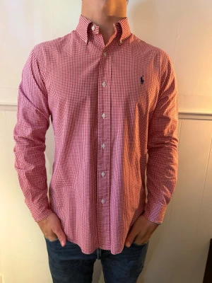 Rödvitrutig skjorta från Ralph Lauren  - Klassisk röd och vit rutig skjorta från Polo Ralph Lauren med button-down krage och broderad logga på bröstet. Skjortan är tillverkad i mjuk bomull. Perfekt nu under sommaren med de röda och vita som bjuder på en ljus skön look. Passar till både kavaj men även till att bara hänga en tröja över axlarna. Skjortan är i fläckfritt skick och har inte några defekter.