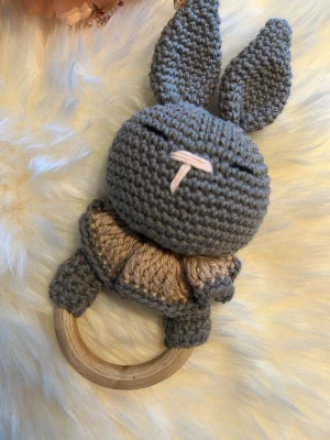 Virkad kanin skallra med träring - Söt handvirkad skallra i form av en grå kanin med långa öron och broderad nos. Kroppen är beige och grå, och skallran har en slät träring som handtag. Perfekt som leksak eller present till små barn.