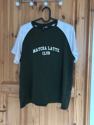 Grön och vit t-shirt Matcha Latte Club - Snygg t-shirt med texten 'MATCHA LATTE CLUB' på bröstet. Tröjan har mörkgrön kropp och vita raglanärmar. Klassisk rund halsringning och avslappnad passform. Perfekt för dig som gillar trendiga och bekväma plagg.