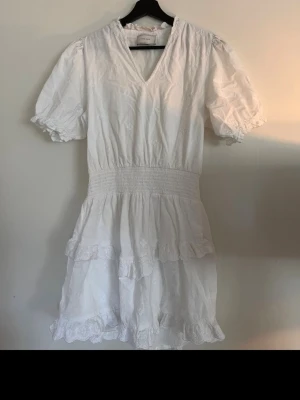 Vit sommarklänning från Neo Noir - Superfin vit klänning från Neo Noir med broderade detaljer, v-ringning och puffärmar. Klänningen har smock i midjan och volanger nertill. Perfekt för varma dagar och har en romantisk vibe. Materialet känns lätt och luftigt.