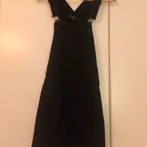 Hängselklänning - Svart, kort hängselklänning i jeansmaterial från Monki. Lös passform. Använd men i mycket gott skick.