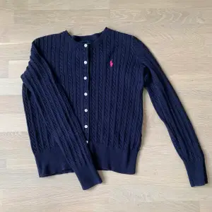 Mörkblå kabelstickad långärmad kofta/tröja från Ralph Lauren. Rosa Ralph Lauren märke på tröjan. Köpt i USA. Nyskick, använd fåtal gånger. Passar för xs.
