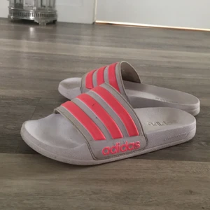 Adidas Tofflor  - Vita Adidas tofflor med rosa ränder💘 Storlek 38 men skulle säga att det är mer som 37. Använda några gånger, (köparen får dom nytvättade) Säljes pga för liten storlek. Nypris: 200kr Mitt pris: 75 först till kvarn! + köparen står för frakten🛍✨