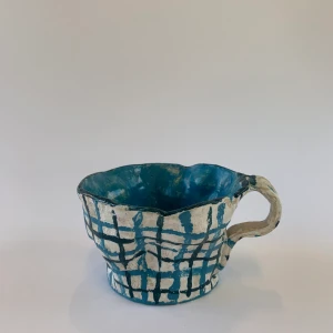 Handgjord vit kopp med blåa rutor - En handgjord kopp i keramik. Koppens yttre har en vit bakgrund med blåa rutor, koppen har även en blå insida. I koppen finns en skåra som är perfekt utformad för bland annat en sked eller tepåse. 