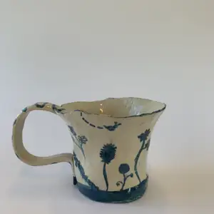 En handgjord kopp i keramik med en naturlig sandfärg samt blåa blommor. 