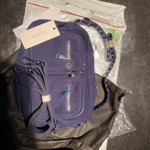 Noella väska i färgen Navy - Helt oanvänd väska ifrån Noella i stilen ”Celina crossover bag”. Väskan är i färgen ”navy” och materialet är mocka imitation. Väskan har kvar både lappar och påsen den kom i. Du får med två band varav ett lite kortare som sitter fast i väskan, och ett längre justerbart band som du kan knäppa loss/på. Väskan har ett stort fack och tre mindre inuti varav ett av dem facken har dragkedja. Väskan har även fack på utsidan. Nypris: ca 700