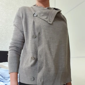 Mångsidig kofta från COS - Kofta från COS i 100% ull, dock ej stickig. Känns som merinoull men det står inte att det är det. Koftan är användbar på många sätt då den har knappar dels i kragen och hela vägen ner på vänster sida, du kan knäppa den på olika sätt och på så sätt förändra utseendet. Färgen är svår att definiera; gråbeige kanske. Använd väldigt sparsamt, finns inget att anmärka på.