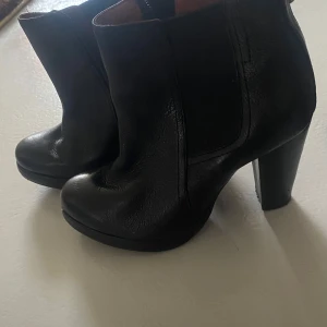 Svarta skinnstövletter med klack - Snygga svarta stövletter i skinn med rund tå och d på sidan. De har en stabil klack som ger extra höjd och stil. Perfekta för att komplettera din outfit med en elegant .