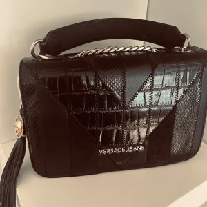 Svart axelväska från Versace Jeans - Snygg svart axelväska från Versace Jeans med ett elegant mönster och kedjedetaljer. Väskan har en praktisk dragkedja och en stilren design som passar perfekt för olika tillfällen. Den har också en dekorativ tofs på sidan.