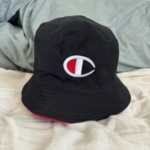 Svart och röd bucket hat från Champion - Snygg vändbar bucket hat från Champion i svart och rött. Ena sidan är svart med Champion-loggan i vitt och rött, medan den andra sidan är röd med Champion-text i blått. Perfekt för att variera din stil!