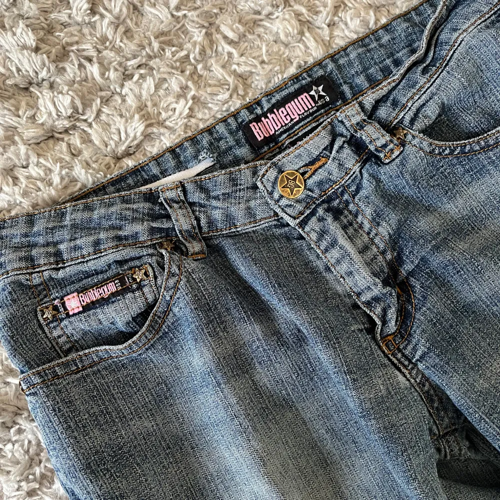 Snygga blå jeans från Bubblegum med klassisk femficksdesign och en stjärnformad knappdetalj. Fina rosa detaljer på. Innerbenslängd: 78 cm. Midjemått rakt över: ca 37. Midjehöjd: 19 cm. Perfekt lågmidjade. Lite slitna längst ned. Kan skicka bild på hela privat, fick inte plats i bilderna här. Farkut & Housut.