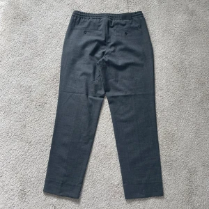 Grå kostymbyxor från J. Lindeberg - Snygga grå kostymbyxor från J. Lindeberg i en ljusgrå melange. Byxorna har en elastisk midja för extra komfort och en stilren design. Perfekta för en elegant look.