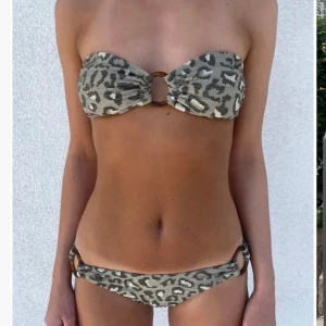 Leopardmönstrad bikini med ringdetalj (oanvänd)  - Trendig bikini med leopardmönster och en snygg ringdetalj framtill. Överdelen är axelbandslös och underdelen har en klassisk passform. Perfekt för en dag på stranden eller vid poolen.