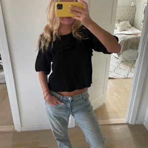 Svart blus med volangdetaljer - Säljer en svart blus med volangdetaljer och korta ärmar. Blusen har en texturerad yta och en lös passform som ger en avslappnad look. Perfekt för att styla med jeans eller kjol.