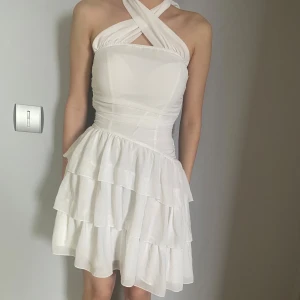 Vit axelbandslös klänning - Säljer en söt vit axelbandslös klänning med volanger och knytband i ryggen. Perfekt för sommarens alla tillfällen. Klänningen har en smickrande passform och är gjord i ett lätt och luftigt material.