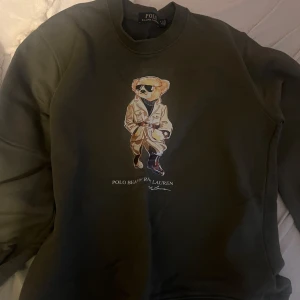 Grön sweatshirt från Polo Ralph Lauren - Säljer en grön sweatshirt från Polo Ralph Lauren med en cool björn i solglasögon tryckt på framsidan. Tröjan har rund hals och långa ärmar, perfekt för en avslappnad stil. Passar bra till jeans eller chinos för en casual look.