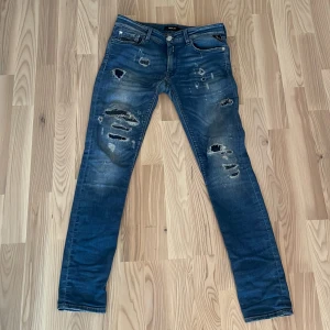 Blå jeans från Replay - Snygga blå jeans från Replay med slitna detaljer och en klassisk femficksdesign. De har en knappgylf och är perfekta för en avslappnad stil. Märkeslogga synlig på knappen och insidan av midjan.