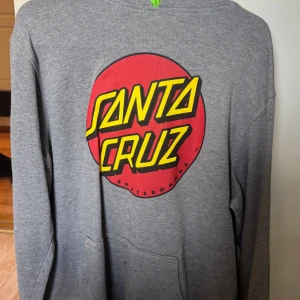 Grå hoodie från Santa Cruz - Säljer en grå hoodie från Santa Cruz med ett stort rött och gult tryck på framsidan. Tröjan har en klassisk känguruficka och långa ärmar. Perfekt för en avslappnad stil.