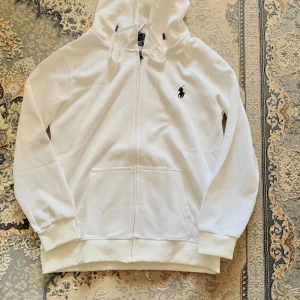 Vit hoodie från Polo Ralph Lauren - Säljer en stilren vit hoodie från Polo Ralph Lauren med dragkedja och det klassiska logotypbroderiet på bröstet. Tröjan har långa ärmar och en justerbar huva med snörning. Perfekt för en avslappnad look.           ✅ Storlek: Storlek M men passar S bättre ✅ Material: Bomullsblandning ✅ Färg: Vit  ✅ Skick: Helt ny, aldrig använd ✅ Pris: 700 Finns för upphämtning i Stockholm eller kan fraktas som tar 1-2 dagar🚚.   Om du är intresserad eller har några frågor, tveka inte att skicka ett meddelande här