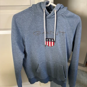 Blå hoodie från GANT - Säljer en snygg blå hoodie från GANT med ett broderat märke på bröstet. Tröjan har en klassisk känguruficka och justerbar huva med snören. Perfekt för en avslappnad stil.