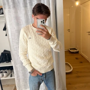 Vit stickad tröja från GANT - Säljer en stilren vit stickad tröja från GANT med kabelstickat mönster. Tröjan har en rund halsringning och ett broderat GANT-emblem på bröstet. Perfekt för en klassisk och tidlös look. 🧶