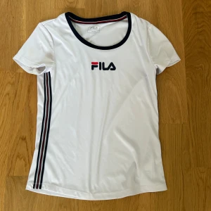 Vit träningströja från FILA - Säljer en snygg vit tränings tröja från FILA med korta ärmar och stilrena ränder i svart och rött längs sidan. Perfekt för en sportig look eller en avslappnad dag. T-shirten har en rund halsringning med kontrasterande kant.