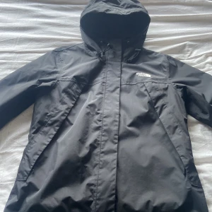 Svart vindjacka från The North Face - Säljer en svart vindjacka från The North Face med huva och dragkedja. Jackan har en stilren design och är perfekt för blåsiga dagar. Den har långa ärmar och logotypen syns både fram och bak.