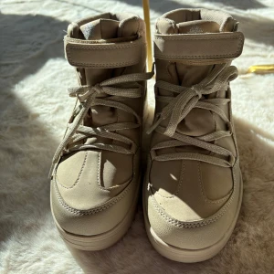 Beige boots med snörning och kardborreband - Säljer ett par beige boots med snörning och kardborreband. Skorna har en rund tå och är tillverkade i syntetmaterial. Perfekta för kyligare dagar med sin robusta design.