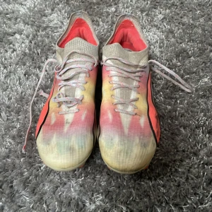 Färgglada fotbollsskor från Puma - Säljer ett par färgglada fotbollsskor från Puma med snörning. Skorna har en mix av beige, rosa och gul färg med en svart detalj på sidan. Perfekta för fotbollsplanen!