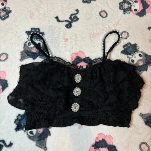 Svart spetsprydd topp från Rojita - Säljer en elegant svart topp från Rojita med spetsdetaljer och pärlprydda axelband. Toppen har volanger och dekorativa knappar framtill. Perfekt för att style ovanpå blusar och toppar 