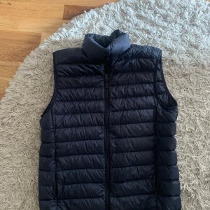 Mörk blå dunväst från Uniqlo  - Snygg mörkblå dunväst från Uniqlo med dragkedja framtill. Perfekt för lager-på-lager under kyliga dagar. Västen har en quiltad design och hög krage för extra värme.