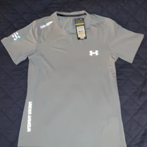 Grå t-shirt från Under Armour - Säljer en grå t-shirt från Under Armour med reflekterande logga och textdetaljer. Perfekt för träning med sin lätta och bekväma passform. Kortärmad design som andas bra.(A-Cop)