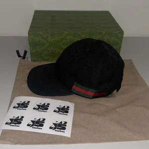 Svart keps från Gucci Canvas  - Snygg svart keps från Gucci med diskret mönster och klassiska gröna och röda ränder på sidan. Perfekt för att ge din outfit en lyxig touch. Finns i S M L