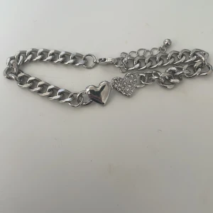 Silverfärgat armband med hjärtan - Snyggt silverfärgat armband med två hjärtan, varav ett är dekorerat med glittrande stenar. Perfekt för att ge en elegant touch till din outfit. Justerbar längd för bästa passform.
