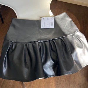 Svart skinnkjol med volang från Even & ODD - Snygg svart skinnkjol med volangdetalj. Perfekt för en trendig look. Kjolen har en kort längd och en stilren design som passar till många olika outfits. Aldrig använt. 