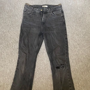 Svarta jeans med slitning - Snygga svarta jeans med en cool slitning på ena benet. De har en klassisk femficksdesign och knappgylf. Perfekta för en avslappnad stil.