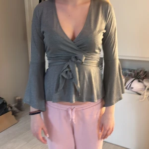 Grå omlottopp med vida ärmar - Snygg grå omlottopp med vida ärmar och knytband i midjan. Toppen har en v-ringad design och är perfekt för en stilren look. Materialet är lite glittrigt