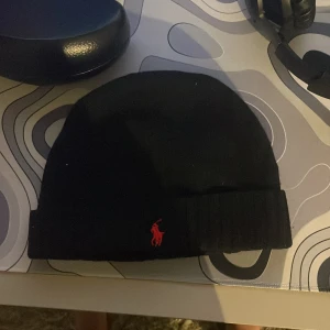 Svart mössa från Polo Ralph Lauren - Snygg svart mössa från Polo Ralph Lauren med det ikoniska röda broderade logotypen framtill. Tillverkad i mjukt material för en bekväm passform. Perfekt för att hålla värmen under kyliga dagar.