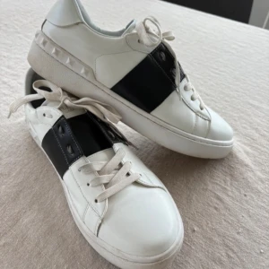 Valentino open - Snygga vita sneakers med en bred svart rem över mitten och dekorativa nitar på sidorna. Skorna har snörning och en rund tå, vilket ger en klassisk look. Perfekta för en stilren och modern stil.
