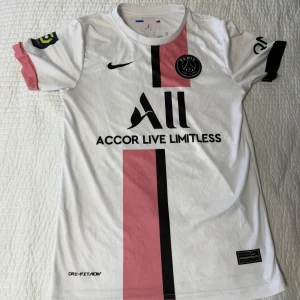 Vit PSG fotbollströja med rosa och svarta detaljer - Säljer en vit PSG fotbollströja från Nike med rosa och svarta detaljer. Tröjan har korta ärmar och texten 'Accor Live Limitless' på framsidan. På baksidan finns namnet 'Sergio Ramos' och nummer 4. Perfekt för fotbollsfans!