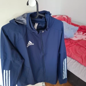 Blå vindjacka från Adidas - Säljer en snygg blå vindjacka från Adidas med dragkedja och huva. Jackan har de klassiska tre vita ränderna längs ärmarna och Adidas-loggan på bröstet. Perfekt för blåsiga dagar!