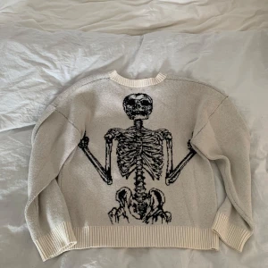 Vit stickad tröja med skelettmotiv (HCW) - Säljer en beige stickad tröja med ett coolt skelettmotiv på baksidan och texten 'HCW' på framsidan. Tröjan har långa ärmar och ribbade muddar vid ärmslut och nederkant. Perfekt för en unik stil! (Kan gå ner i pris)