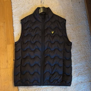 Dunväst lyle and scott - Snygg svart dunväst från Lyle & Scott med vågigt quiltat mönster och dragkedja framtill. Den har en gul broderad logotyp på bröstet. Perfekt för lager-på-lager under kyliga dagar. Jag har aldrig använt 