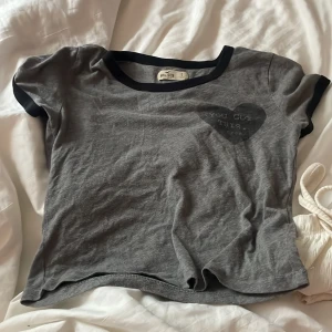 Grå t-shirt från Hollister - Säljer en grå t-shirt från Hollister med svart kant vid hals och ärmar. T-shirten har ett hjärta med texten 'You Got This' på bröstet. Perfekt för en avslappnad stil.