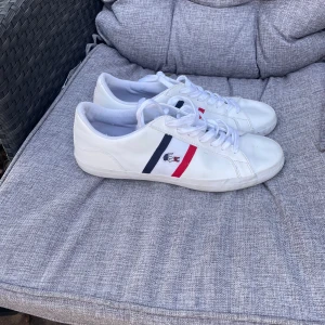 Vita sneakers från Lacoste - Snygga vita sneakers från Lacoste med klassiska ränder i blått och rött på sidan. Skorna har snörning och en rund tå, vilket ger en stilren look. Perfekta för en avslappnad stil.