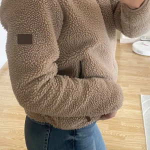 Beige teddyjacka med dragkedja - Mysig beige teddyjacka med dragkedja framtill. Perfekt för kyliga dagar med sin fluffiga och varma design. Jackan har en normal passform och långa ärmar.