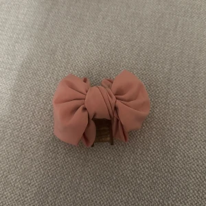 Rosa hårklämma med rosett - Söt rosa hårklämma med en stor rosett. Perfekt för att ge en extra touch till din frisyr. Klämman är gjord av plast och har en mjuk, tygklädd yta.
